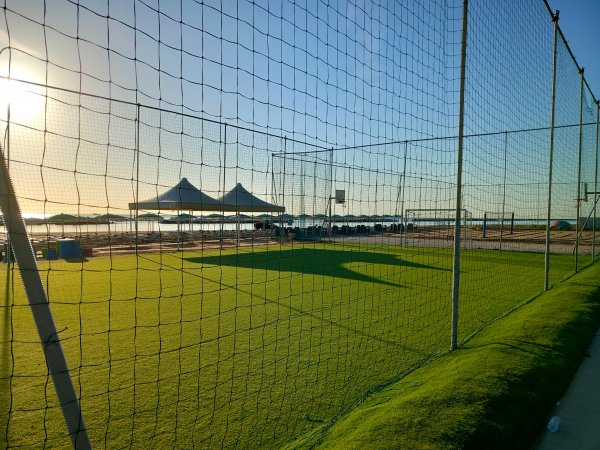 Campo da calcio in erba con rete, tende in lontananza e mare al tramonto.