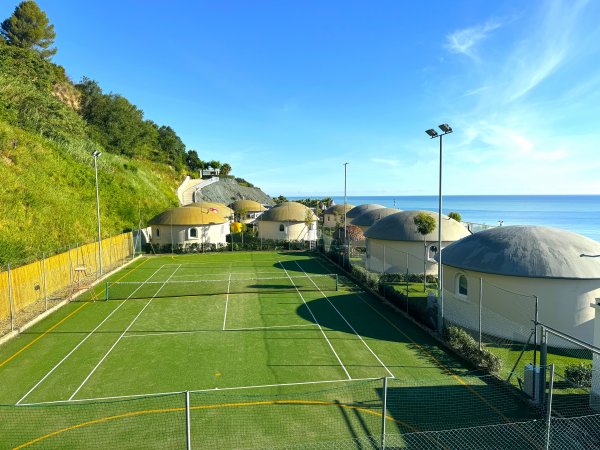 Campo da tennis in erba, circondato da strutture rotonde e vegetazione con vista mare.