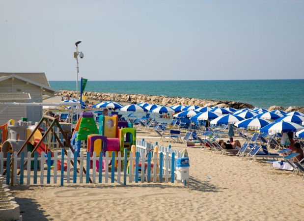 Spiaggia con ombrelloni blu e bianchi, area giochi recintata e mare calmo.