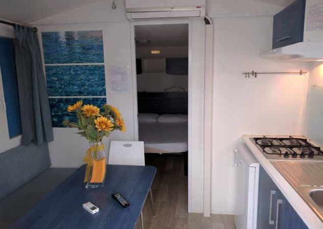 Interno di un bungalow con cucina, tavolo blu, sedia, e un vaso di fiori gialli su un tavolo.