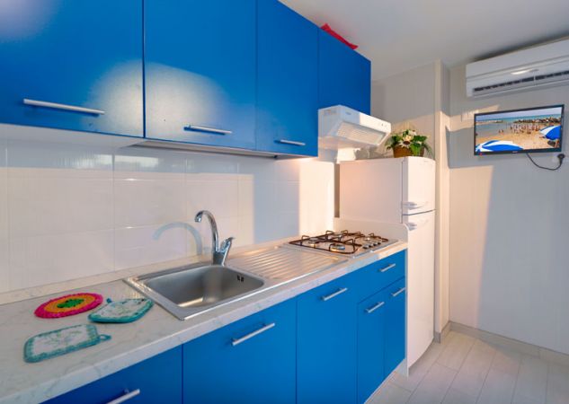 Cucina con mobili blu, lavello inox, fornello a gas, frigo bianco, aria condizionata e TV.
