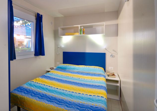 Camera con letto matrimoniale, copriletto a strisce gialle e blu, mensole e finestre.