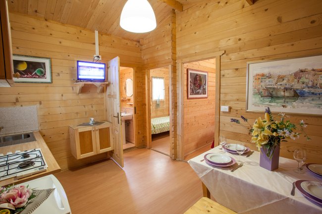 Cucina in legno chiaro con tavolo apparecchiato, porta aperta sul bagno, TV sullo scaffale.
