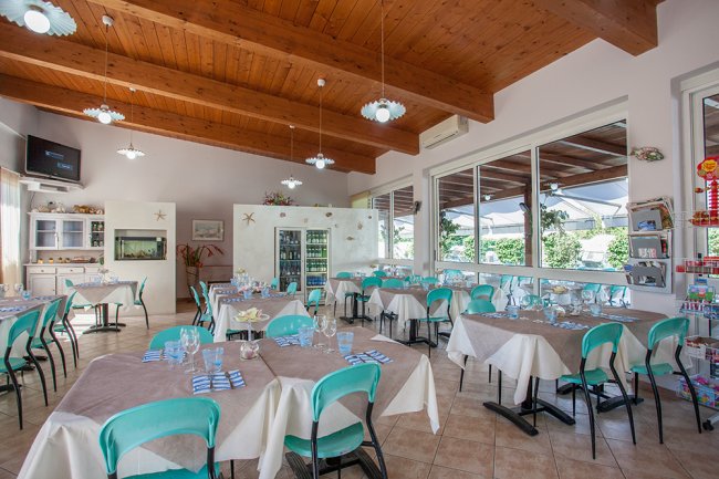 Sala ristorante luminosa con tavoli apparecchiati e sedie turchesi, soffitto in legno.
