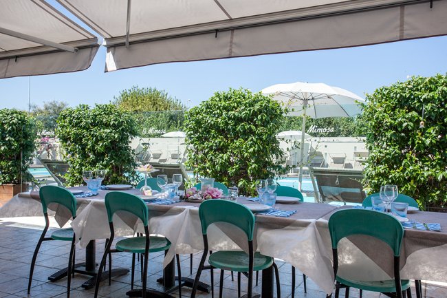 Tavola apparecchiata con sedie verdi sotto un gazebo con piante e piscina sullo sfondo.