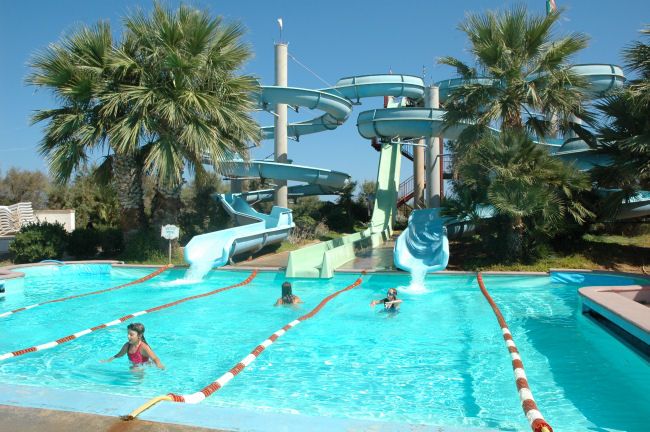 Piscina con scivoli d'acqua e ragazzi che nuotano, circondata da palme.