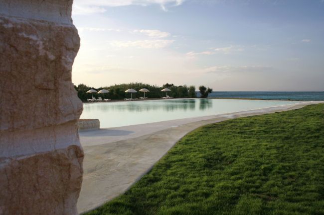 Piscina vista mare, contornata da prato verde e ombrelloni bianchi.