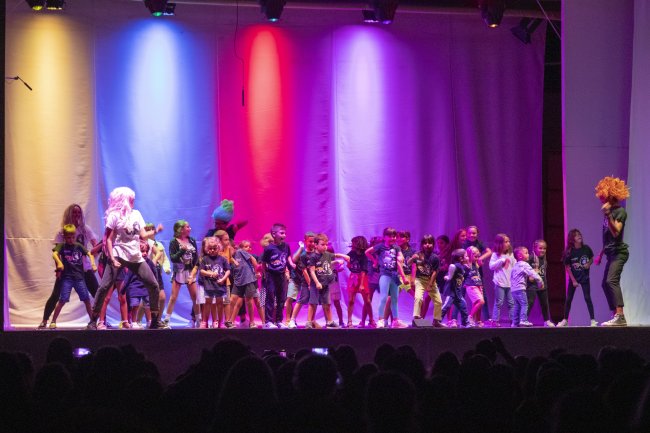 Gruppo numeroso di bambini su un palco colorato durante una performance teatrale o musicale.