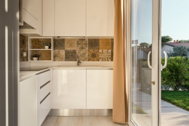 Cucina moderna con mobili bianchi, piastrelle decorative e porta-finestra aperta verso il giardino.