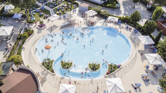 Piscina affollata con bambini che giocano, circondata da ombrelloni bianchi e area verde.