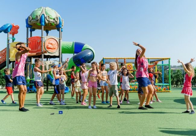 Bambini e animatori danzano allegri in un parco giochi all'aperto con scivoli colorati.
