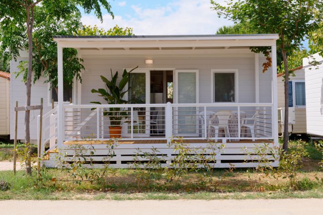 Casa mobile bianca con veranda, sedie e pianta in vaso davanti.