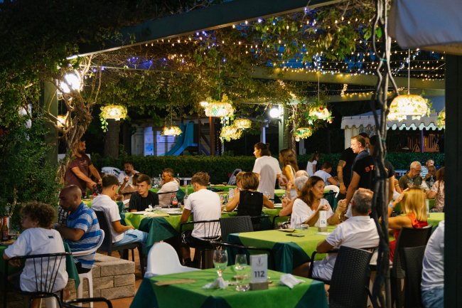 Persone sedute a tavoli verdi all'aperto in un ristorante, illuminato da luci e lanterne.