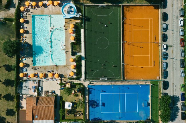 Vista aerea di una piscina, un campo da calcetto e due campi da tennis con ombrelloni gialli.
