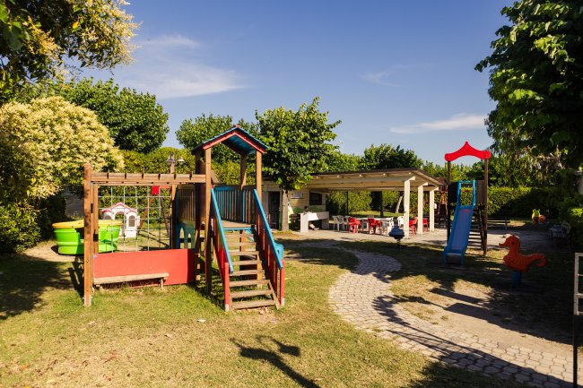 Un parco giochi per bambini con scivolo, altalena e aree per sedersi, circondato da alberi.