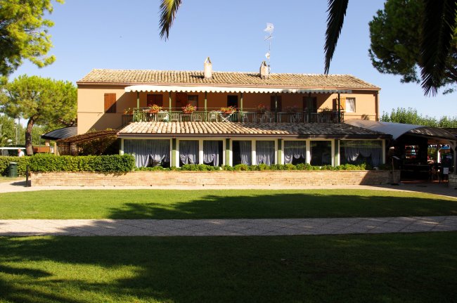 Una casa colonica a due piani con veranda e balcone, circondata da un prato e alberi.