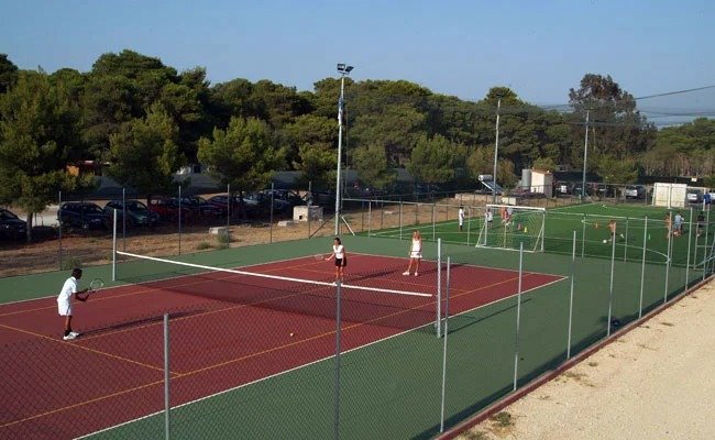 Campo da tennis all'aperto con persone che giocano, circondato da alberi e recinzioni.