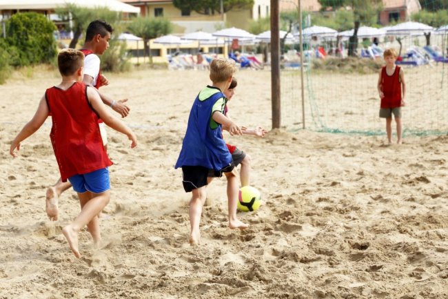 Bambini in maglie colorate giocano a calcio sulla sabbia vicino a una porta improvvisata.