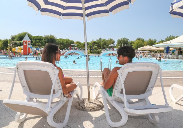Due persone su sdraio sotto un ombrellone a bordo piscina, con giochi acquatici sullo sfondo.
