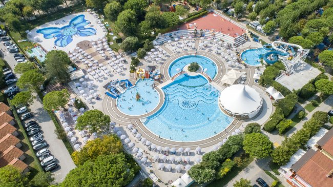 Complesso acquatico con piscine, ombrelloni e scivoli, circondato da alberi e un'area giochi.