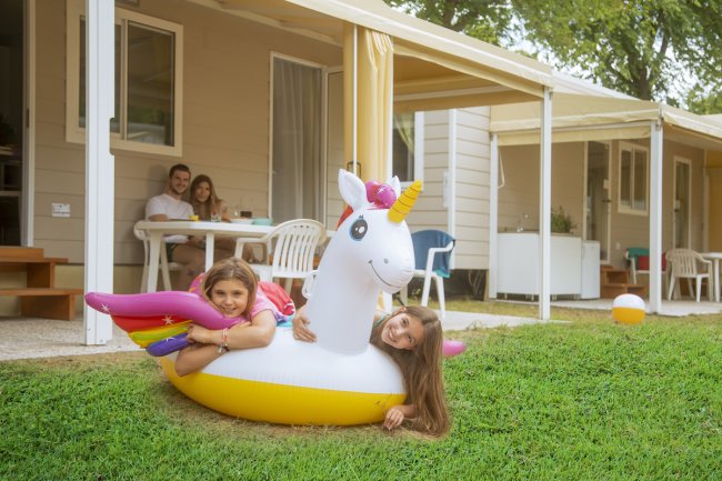 Bambine con gonfiabile unicorno su prato, famiglia a tavola sullo sfondo.